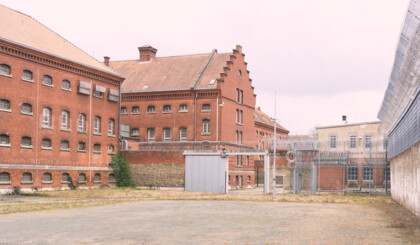 Ein Bild der JVA Rennelberg. Ein rotes Backsteingebäude mit vergitterten Fenstern, einen Hof mit Zäunen und Stacheldraht unter grauem Himmel.